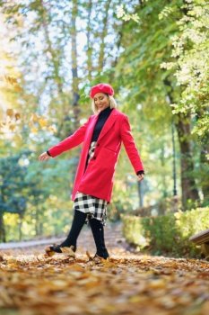 Full body of positive female in red clothes and beret walking on pathway with withered leaves in autumn park with trees. Fashionable woman on path with fallen leaves