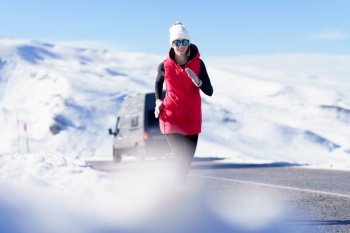 Smiling female wearing warm vest and hat with sunglasses jogging on road in amazing mountains in winter. Sportive woman in warm clothes running on road in mountains