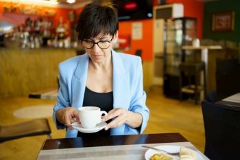 Concentrated middle age female in formal wear and eyeglasses, sitting at table with cup of coffee in hands while eating and looking down in modern cafe. Pensive woman with cup of coffee in cafe