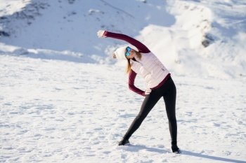 Full body of young female in cap polarized sunglasses, looking away and stretching arm sideways while standing with legs apart and hand on thigh on snowy mountain in daylight. Young woman standing and doing exercise on snow in highlands