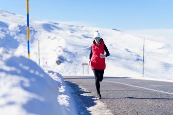 Full body of woman in outerwear and sunglasses jogging on asphalt road with snowy mountains. Female running on road in snowy mountains