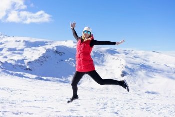 Full body of excited young female in vest cap, polarized sunglasses looking at camera while leaping up in air with stretched arms against snowy mountains and blue sky in daylight. Carefree young woman jumping and enjoying vacation on snowy slope