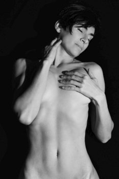 Black and white of sensual adult female with short dark hair covering naked breast and touching neck with closed eyes in studio. Sexual naked woman touching neck with closed eyes in studio