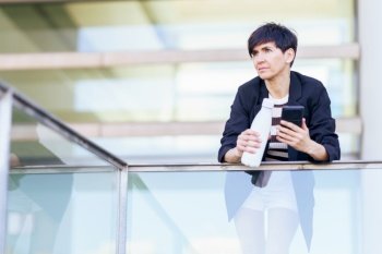 Pensive female in black coat with short hair and smartphone looking away while leaning on glass fence of modern building and holding bottle. Women with smartphone and white bottle of water