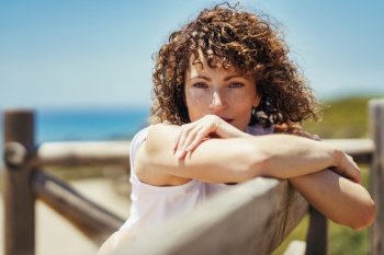 Calm young female with curly hair in white outfit leaning against fence and looking at camera while relaxing alone near blurred background of sea. Young woman leaning on wooden handrail at seaside
