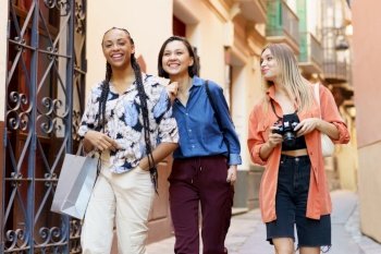 Cheerful young multiracial women in trendy clothes laughing while passing by building wall on blurred background. Smiling female diverse friends having conversation near building
