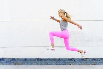 Full body side view of female athlete in pink leggings and white sneakers, looking away while jogging during running exercise on street next to shabby rusting iron ladder in daylight. Woman in sportswear running on street in daylight