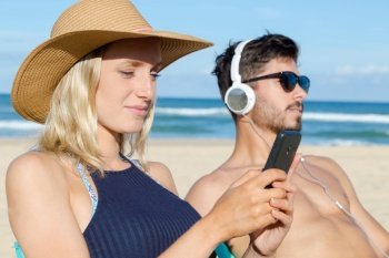 couple on beach man listening to headphones woman using smartphone