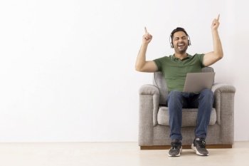 Cheerful young man listening music on headphones and having fun with raising his  hands while sitting on sofa