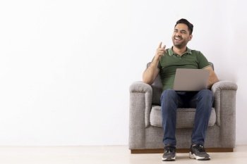 Portrait of a cheerful young man pointing elsewhere while sitting on sofa with laptop on his lap