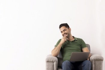 Cheerful young man with hand on chin looking away while sitting on sofa with laptop on his lap