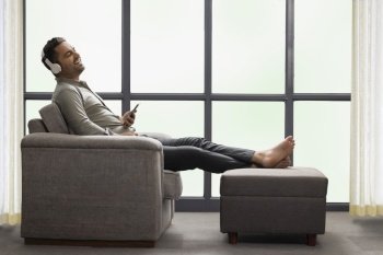 Young businessman listening music on headphone using mobile phone while leaning on sofa at home