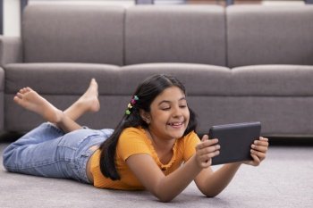 Portrait of a cheerful girl using digital tablet while lying down on floor in living room 