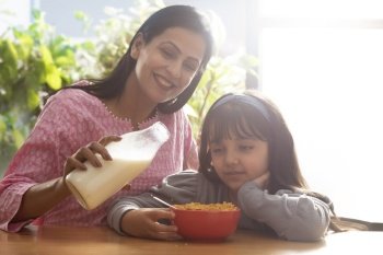 A HAPPY MOTHER POURING MILK INTO A BOWL OF CORNFLAKES FOR DAUGHTER