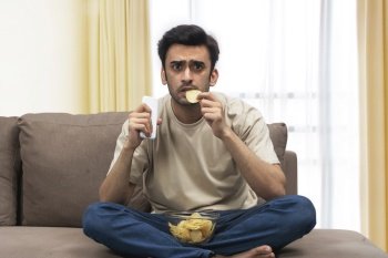 A young man with remote sitting tensed on sofa eating chips while watching sports.