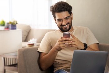 A man sitting with laptop smiling while working on his mobile.