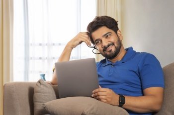 A man sittting on couch with laptop daydreaming while holding specs in hand.