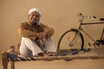 A RURAL MAN COMFORTABLY SITTING ON COT AND LOOKING AT CAMERA