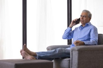 Serious senior man talking on mobile phone while sitting on sofa at home