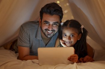 A FATHER AND DAUGHTER LOOKING AT CAMERA WHILE USING DIGITAL TABLET AT NIGHT