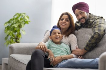 A SIKH BOY SMILING AND LEANING OVER MOTHER SITTING ON SOFA WHILE FATHER BENDS OVER TO THEM AND SMILES