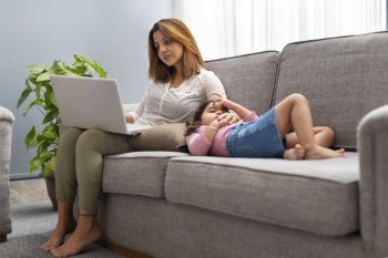 A WOMAN WORKING ON HER LAPTOP WHILE PATTING DAUGHTER TO SLEEP ON SOFA