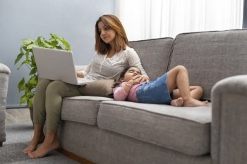 A WOMAN WORKING ON LAPTOP WHILE DAUGHTER COMFORTABLY SLEEPING NEAR HER ON SOFA
