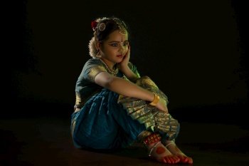 Young bharatnatyam dancer sitting during her performance in front of a black background. 