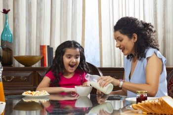 Mother pouring milk into bowl with chocos for her daughter during breakfast at home