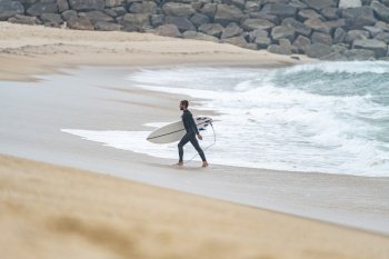Side view of man with surfboard on beach. Side view of surfer in wetsuit holding board and walking from sea waves. Surfing concept.