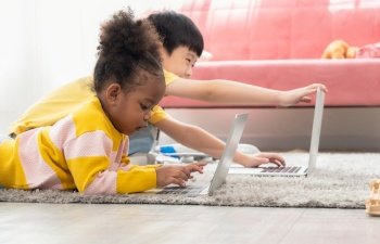 Happy black little curly girl and asian boy in yellow shirt using laptop at home. Two child lying on carpet in living room.