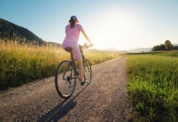 Woman is riding a mountain bike on gravel road at sunset in summer. Colorful landscape with sporty girl riding bicycle, field, dirt road, green grass, blue sky. Sport and travel