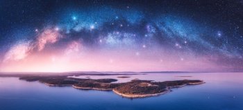 Milky Way arch and islands in the sea at summer night. Kamenjak cape, Adriatic sea, Croatia. Landscape with purple starry sky, arched milky way, sea coast, water, mountains. Top view. Panorama