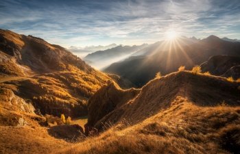 Mountains in low clouds at beautiful sunset in autumn in Dolomites, Italy. Landscape with alpine mountains, orange grass and trees, hills in fog, blue sky with clouds and sun in fall. Nature