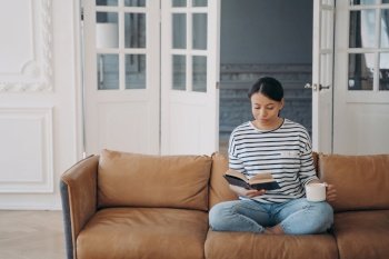 Woman is reading novel and drinks coffee or tea. Happy young european woman is reading book sitting on comfortable leather couch. Lady enjoying poetry. Living room interior with copy space.. Woman is reading novel and drinks coffee sitting on comfortable leather couch. Lady enjoying poetry.