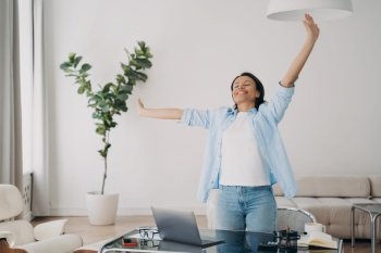 Happy female office worker, standing stretching at workplace feels free from life’s problems. Smiling pleased woman relaxing enjoying break after successful done work at desk with laptop.. Happy female stands stretching at workplace, relaxing enjoying break after successful work at laptop