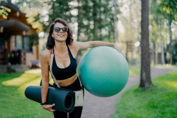 Joyful female smiles broadly, holds big fitness ball, rolled up karemat, wears sunglasses, enjoys summer day, goes in for sport regularly, being pilates instructor, poses outdoor at beautiful nature