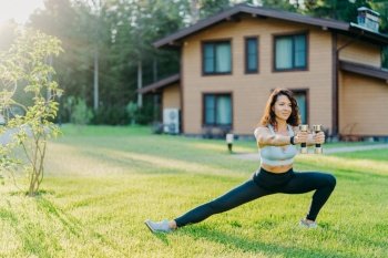 European sportswoman holds dumbbells and stretches legs, dressed in sport clothes, does physical exercises outdoor near house early in morning. Healthy lifestyle, weightlifting and fitness concept