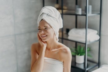Positive European woman smiles pleasantly, touches healthy skin, wears wrapped towel on head, poses in bathroom. Spa lady with natural makeup poses refreshed after taking shower. Pampering, wellness