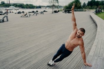 Self determined muscular man dressed in sport clothes stands in side plank, keeps arm raised, has workout outdoor during summer day. Sport, healthy lifestyle, strength and endurance concept.