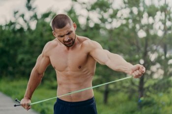 Young handsome strong European man poses at park stadium, stretches skipping rope, has naked torso and muscular arms, going to have physical exercises, poses in open air. Street workout concept