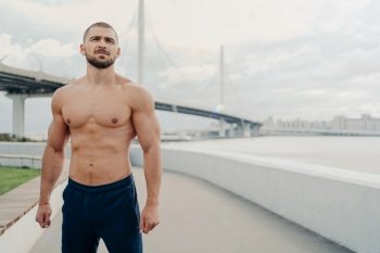 Handsome muscular man with naked torso has outdoor fitness workout looks into distance, has sexy body, dressed in sports trousers stands outside near river bridge. Athletic guy runs in morning