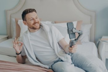 Attractive bearded man in casual clothes lying on bed while chatting online with friends on smartphone, smiling and showing with his hand okey sign, holding cellphone with gimbal stabilizer handheld. Attractive bearded man in casual clothes lying on bed while chatting online