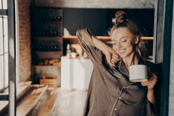 New day. Happy beautiful brunette female stretching with cup of coffee in her hand enjoying morning sunshine while standing in kitchen doorway in satin pajama. People and lifestyle concept. Happy beautiful brunette female stratching with cup of coffee in her hand while standing in kitchen