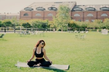 Relaxed slim brunette Caucasian woman sits in lotus pose on fitness mat has perfect body shape does yoga and meditation wears sports clothes poses outdoor. Health fitness and relaxation concept