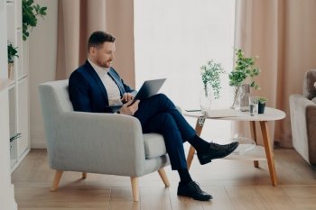 Focused young businessman in stylish suit sitting in armchair in living room at modern apartment and looking at laptop screen while working remotely at home. Business people and remote job. Concentrated man in suit typing on laptop in living room