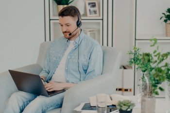 Busy man dressed in denim clothes supplies consultant service types on laptop computer wears headphones with microphone works distantly sits in comfortable armchair participates in online negotiations