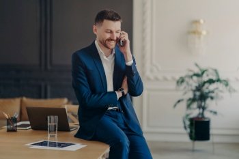 Handsome businessman in formal clothes talking on phone in office and being happy to hear good news about project while sitting on work desk with laptop in luxury apartment or office. Business concept. Smiling executive manager talking on phone while sitting on table in office