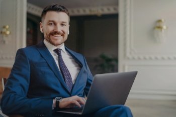 Young successful confident businessman in formal elegant suit working with laptop computer at his workplace in office, smiling at camera and feeling happy. Business occupation concept. Happy man executive manager looking happy and satisfied with business results while working with laptop in office
