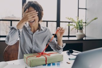 Excited young woman office employee closing eyes with hand unpacking Xmas present pulling ribbon when sitting at workplace, happy freelancer lady opening gift box with excitement during remote work. Excited young woman office employee closing eyes with hand while unpacking Xmas present at work